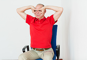 Ein Mann sitzt auf einem Stuhl. Er hat Zeigefinger und Daumen zu Kreisen geschlossen und vor die Augen gelegt.