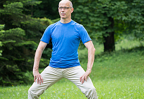 Mann steht mit geradem Oberkörper, gespreizten Beinen und leicht gebeugten Knien; Hände sind auf die Oberschenkel gestützt