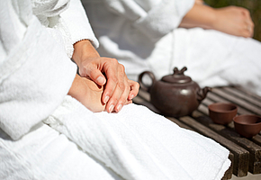 Die Hände einer Frau, die im Bademantel neben einer Teekanne und zwei Bechern sitzt.