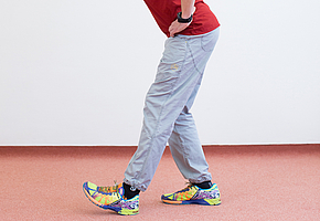 Mann steht im Raum mit leicht nach vorn geneigtem Oberkörper, Hände in die Hüften gestützt; der Fuß des linken Beines ist leicht nach vorn gestellt, die Fußspitze angehoben.